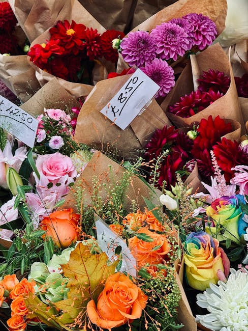 A vibrant assortment of fresh flowers displayed for floral gift or arrangement, including red dahlias, purple chrysanthemums, pink roses, orange spray roses, and pastel rainbow roses, all wrapped in b