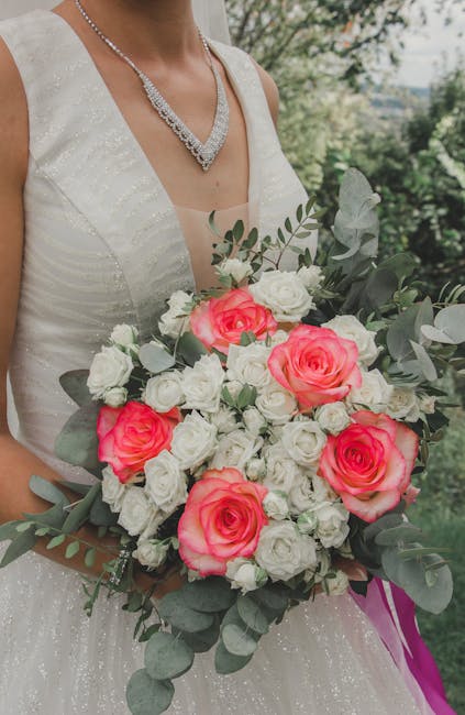 A bride wearing a white, sleeveless wedding gown with subtle shimmer, holding a large, rounded floral bouquet. The bouquet features vibrant pink roses, white roses, and small white filler flowers, acc