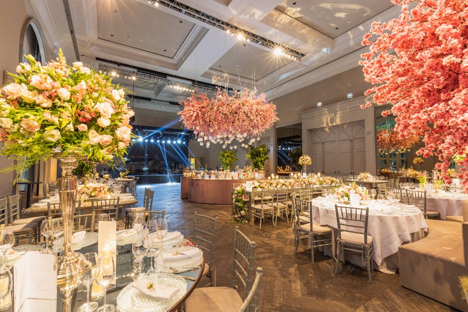 An elegantly decorated banquet hall featuring a floral arrangement for a wedding event, with large bouquets of pale pink roses and white lilies in tall, shiny silver vases placed on round tables, comp