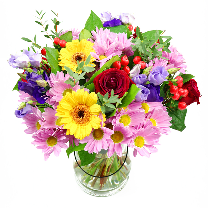 A vibrant, garden-style bouquet is shown standing proudly in a clear glass vase, its colours glowing against a pure white background that makes every bloom pop. The central focus is a cluster of sumptuous deep red roses, their petals plush and velvety, curling inward to create a rich, romantic core. Just beside them, two sunny yellow gerbera daisies open wide, each one a circle of golden petals framing a warm brown-orange centre, like tiny suns shining over the streets of Hounslow on a bright spring morning. Encircling this heart, generous sprays of soft pink daisies or chrysanthemums cascade outward, each flower made of many slender petals and a delicate yellow centre, adding a gentle, reassuring charm and plenty of volume. Nestled between the pinks and yellows are graceful stems of lisianthus in shades of purple and lavender, their bell-shaped blooms slightly ruffled at the edges, adding a sophisticated, almost whimsical touch reminiscent of borders in a well-kept garden near Osterley Park. Tiny clusters of bright red hypericum berries peek out from the greenery, their round, polished surfaces catching the light and bringing a playful, textural accent. The whole bouquet is threaded with layers of foliage: broad, glossy dark green leaves that frame the flowers, and lighter, more delicate green sprigs that arch outward, softening the silhouette and giving the arrangement a fresh, just-picked feel. Through the glass vase, the stems are visible, carefully cut to even lengths and arranged in clear water, hinting at the thoughtful craftsmanship behind the design. The composition feels joyful and generous, perfect for marking birthdays, thank-you gestures, or house-warmings anywhere from Hounslow Central to the quieter residential streets, bringing a burst of colour and life indoors.